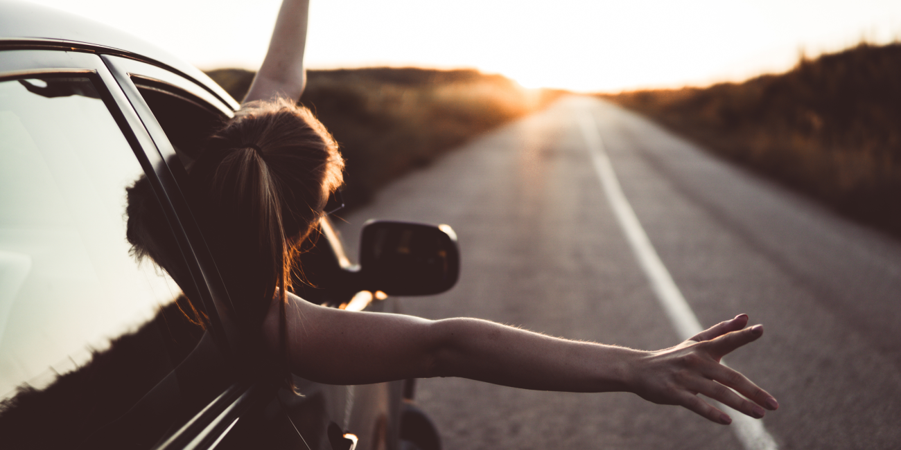 woman with her head and hands leaning out of a car window as it travels down the road