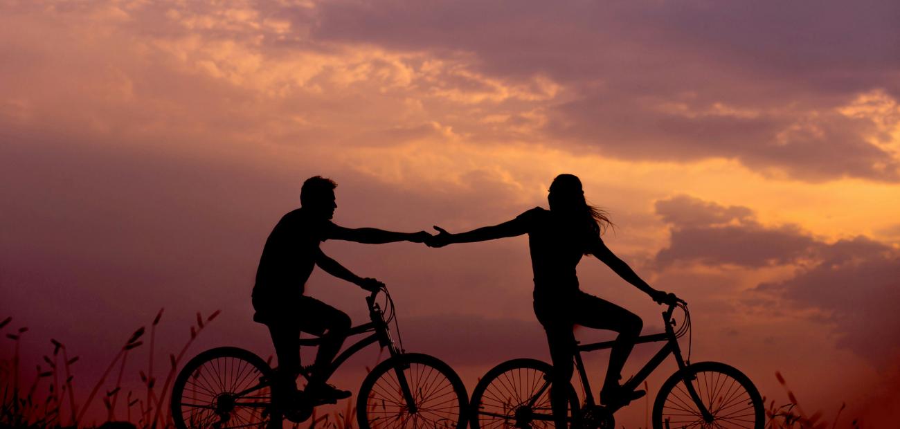 a silhouette of a young couple riding bicycles with their arms out stretched to each other at sunset